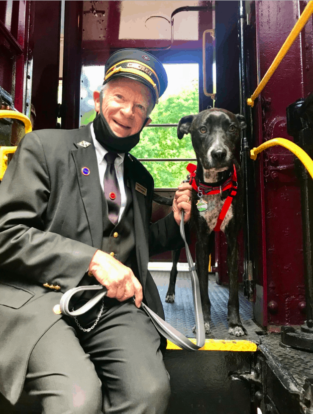 Happy train conductor sitting next to a brindle colored dog in a red harness on the Leigh Gorge Scenic Railway in Jim Thorpe, PA